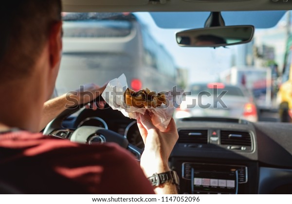 Man driving car while eating hamburger. Waiting and\
standing in traffic jam