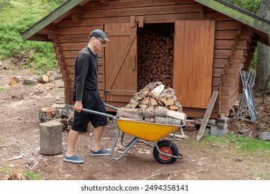 Man driving a wheelbarrow and wearing black clothes in the forest of Finland near the Yellow metal garden wheelbarrow and a pile of sawn firewood near the firewood storage. Preparing firewood 