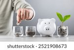 Man drops coins into a glass jar, with stacks of coins and a wooden house model beside him. Emphasizes the concept of saving money for future house investments and financial stability