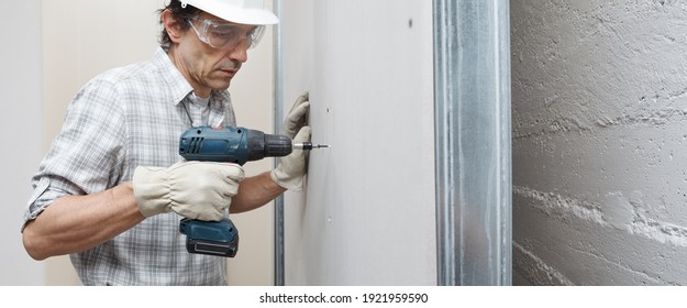 man drywall worker or plasterer using cordless electric screwdriver to fix the plasterboard sheets to the metal profiles to build the new wall . Wearing white hardhat, work gloves and safety glasses