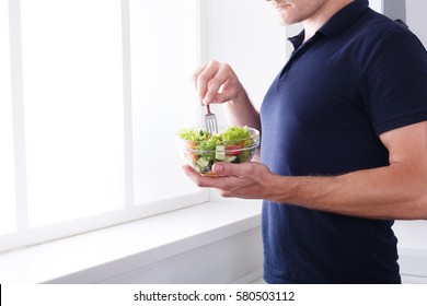 Man Eat Healthy Lunch In Modern Interior. Unrecognizable Profile Male Torso In Blue T-shirt, Hand With Fork, Near Window With Vegetable Salad In Bowl, Diet Food Concept. High Key Image