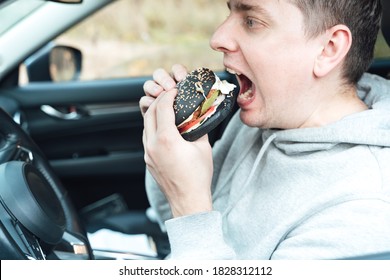 Man Is Eating Black Burger With Meat, Fish, Tomato, Salad, Sause In Car. Fast, Safe, Takeaway Food On The Road To Coronavirus Pandemic. Delicious American Snack. Family Dinner In Trip, Travel.