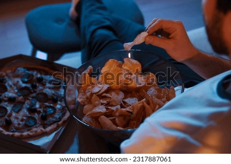 Man eating chips and pizza while watching TV on sofa at night, closeup. Bad habit