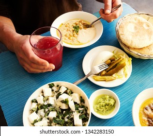 Man Eating Fresh Warm Hummus With Pita Bread, Pickles, Feta Cheese Salad With Organic Herbs And Drinking Fresh Squeezed Fruit Juice. Healthy Eating Middle Eastern Cuisine Background.  Foodie Travel. 