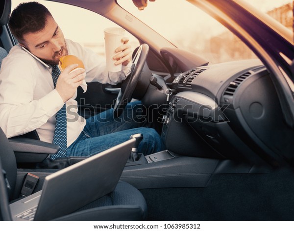 Man\
eating an hamburger and working seated in his\
car