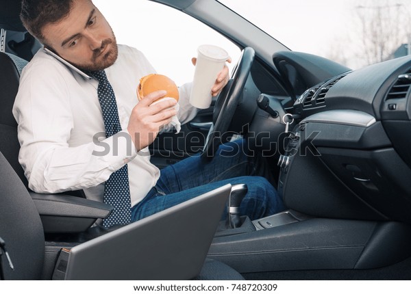 Man\
eating an hamburger and working seated in his\
car