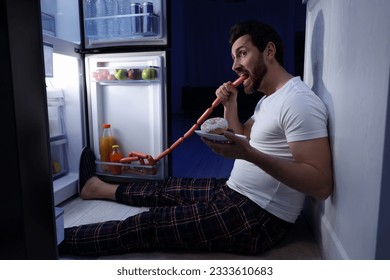 Man eating sausages near refrigerator in kitchen at night. Bad habit