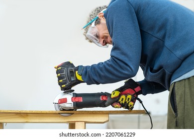 Man With Electric Saw To Cut Wooden Piece At Home. He Is Wearing Protective Glasses And Gloves. Close Up