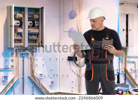 Man electrician. Electrical equipment repair specialist. Guy builder near energy shield. Electrician with wiring repair manual. Electrification industrial buildings. Man electrician with tablet