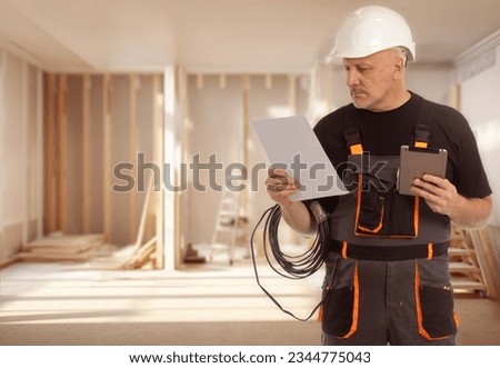 Man electrician. Worker with tablet and papers. Electrician in building under construction. Builder guy. Electrician in room being renovated. Man is wiring master. Electrification specialist