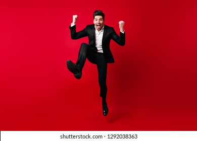 Man In Elegant Suit Fooling Around In Studio. Emotional Male Model Jumping On Red Background.