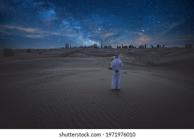 Man With Emirates Outfit Clothes Standing Alone In The Desert During The Night And Watching The Milky Way Galaxy And The Dubai Skyline