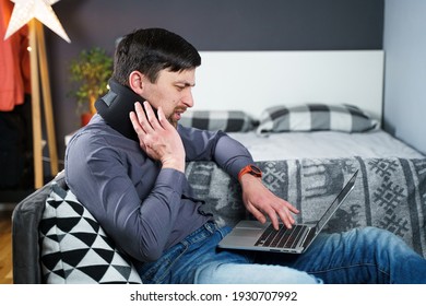 Man Employee After Accident Working At Home Behind Laptop And Feels Pain In Neck, Uses Black Neck Collar Support. Male With Neck Injury Freelance Programmer On Sick Leave Working At Computer On Couch.