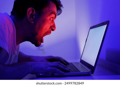 A man is engaged in intense work late at night, leaning toward his laptop. The room is illuminated with a striking blue light that creates a dramatic ambiance. 
A young man is frustrated and angry.