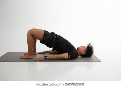 A man is engaging in the bridge exercise on a yoga mat in a well-lit indoor studio. He emphasizes strength and stability as he raises his hips, focusing on core and glute engagement - Powered by Shutterstock