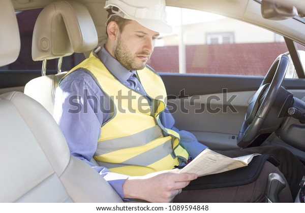 Man engineer builder in a white\
hard hat, shirt and yellow waistcoat sits in the car and\
reads
