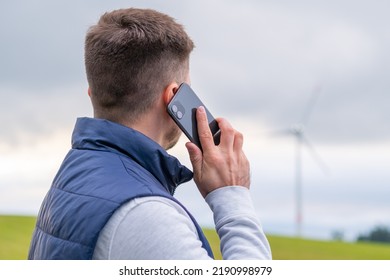 Man engineer reports about functioning wind turbine via smartphone looking at windmill. Wind turbine produces green energy against grey sky