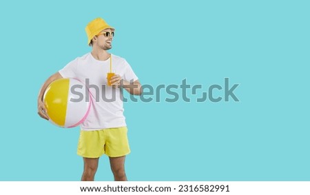 Man enjoying summer holiday. Happy guy wearing white tee shirt, vacation shorts, yellow panama hat and sun glasses standing on blue studio background, holding beach ball and looking at copy space side