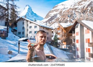 Man Enjoying Wine In Swimming Pool Against Town And Mountains. Tourist Relaxing In Hot Tub Against Majestic Matterhorn. Scenic View Of Snowy Landscape In Alps.