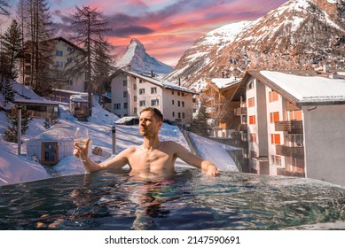 Man Enjoying Wine While Swimming In Pool Against Townscape. Tourist Relaxing In Hot Tub Against Matterhorn Mountain. Scenic View Of Snow Covered Landscape During Sunset.