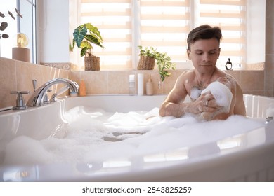 A man enjoys a bubble bath, lathering his arm with soap in a bright, modern bathroom. Surrounded by plants and bath products, the scene conveys self-care, relaxation, and personal hygiene. - Powered by Shutterstock