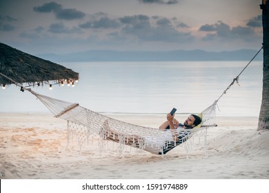 A man enjoys calm, lies in a hammock with a phone on the background of the ocean and sunset. A handsome guy looks, reads on the phone at sunset on a tropical island beach. - Powered by Shutterstock