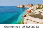 A man enjoys a peaceful moment on a stone ledge, overlooking the vibrant turquoise sea of Dubrovnik. The historic city and boats dot the horizon under a clear blue sky. men on vacation in Croatia