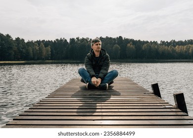 A man enjoys the view while exploring nature while sitting cross-legged on a wooden pier in autumn.