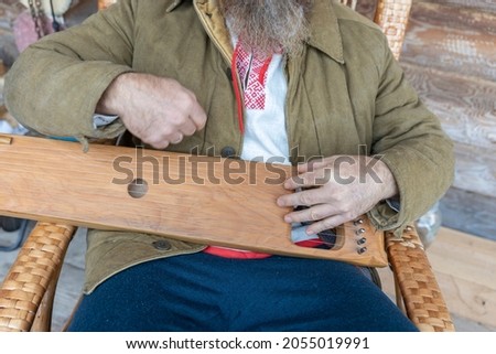A man in ethnic clothes plays an ancient musical instrument. A bearded man, a forester has built a house and is playing a psaltery. 