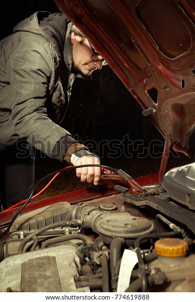 Man in evening winter time chyrging discharged car\
battery with cables