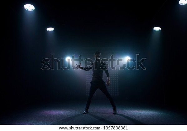 A man\
in everyday clothes stands on a dark stage in a dance pose. Back\
lighting stage light. Silhouette of a\
dancer.