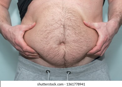 a man with excess weight and a big body holds his stomach with his hands, gray wall background, front view