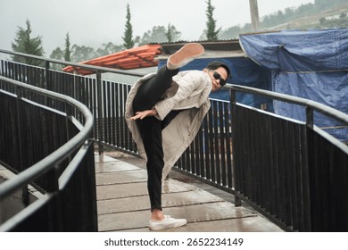 A man executes a high kick while standing on a railing path outdoors, exuding confidence and agility during a rainy day. He wears casual clothing and sunglasses, displaying both style and energy.