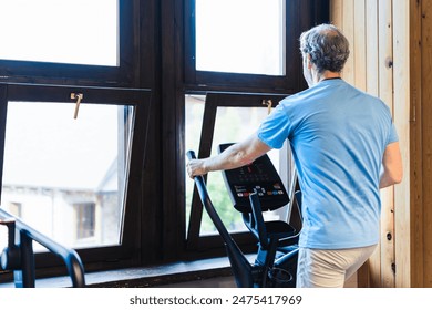 A man is exercising on a stationary bike in front of a window. The man is wearing a blue shirt and white pants - Powered by Shutterstock