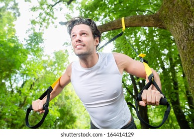 man exercising with suspension trainer sling in City Park under summer trees for sport fitness