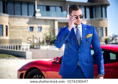 Man in expensive custom tailored suit with glasses posing outdoors in front of expensive car and house