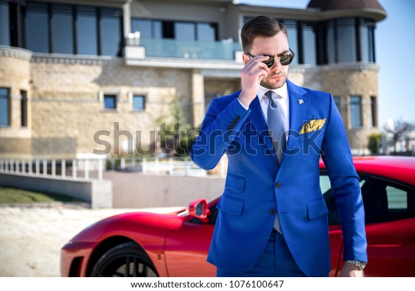 Man in expensive custom\
tailored suit with glasses posing outdoors in front of expensive\
car and house
