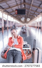 Man experiences boredom and sadness while waiting for his train, resting his head on his hand next to his luggage on a bench at a long, empty station platform