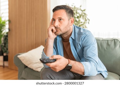 Man experiencing boredom and listlessness while watching entertainment at his residence, feeling disengaged and apathetic towards the content, looking weary with a remote control