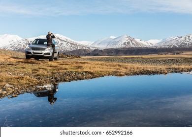 Man Explorer In Iceland Sitting On The Car. Adult Man On The Bonnet Looking Through Binoculars. Majestic Mountains In Background, Reflections In Water On Foreground. Travel And Wanderlust Concept