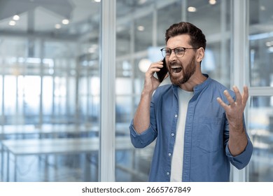 Man expressing frustration while speaking on phone in a modern office setting. A concept of stress, work pressure, or heated discussions in personal or professional life.