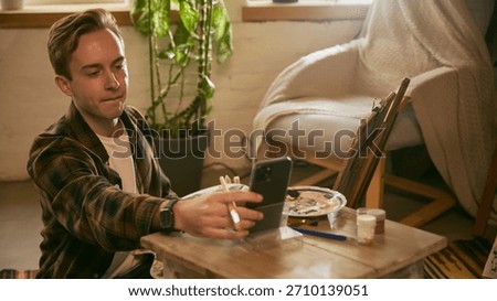 Man extending arm to smartphone while painting. Concept of checking references, consulting tutorials, balancing tools, and maintaining creative flow in a home art setup.