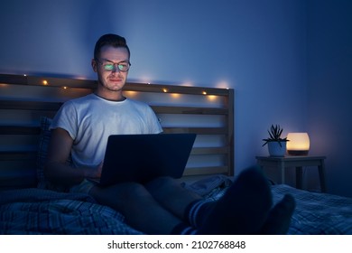 Man with eyeglasses sitting on bed in bedroom and using laptop. Face illuminated from computer display.
