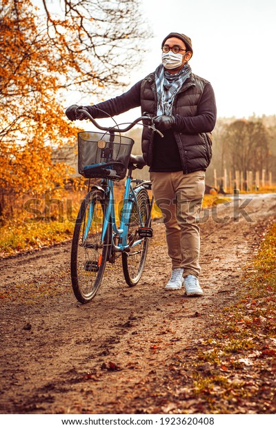 Man with face mask and glasses\
walking with a blue bike outside in autumn a gravel\
path
