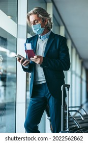 Man With Face Mask Standing At Airport Waiting Area Using His Mobile Phone. Traveler Waiting For His Flight At Airport Departure Area.