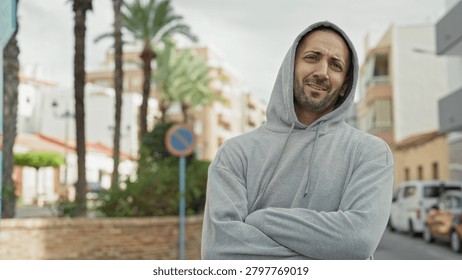 Man with face visible pulls and adjusts hoodie while crossing arms and looking sideways on a palm lined street with parked cars; thoughtful resilience.