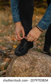 Man In Fashion Clothes Is Tying His Shoelaces On A Rock In An Autumn Park. Guy In Black Leather Sneakers 