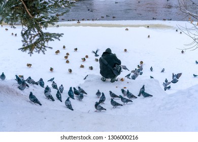 A Man Is Feeding Doves And Ducks On The Frozen River Bank In A Small Old Town In Winter