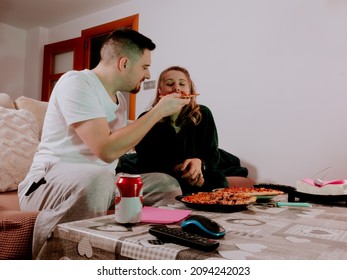 Man feeding pizza to woman sitting on sofa at home next to a table with a remote control and beer