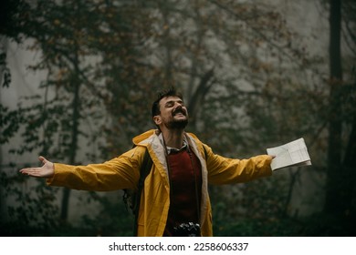 Man feel free concept. Lost male hiker holding a map while standing with arms spread during exploring a national park on a rainy day. - Shutterstock ID 2258606337